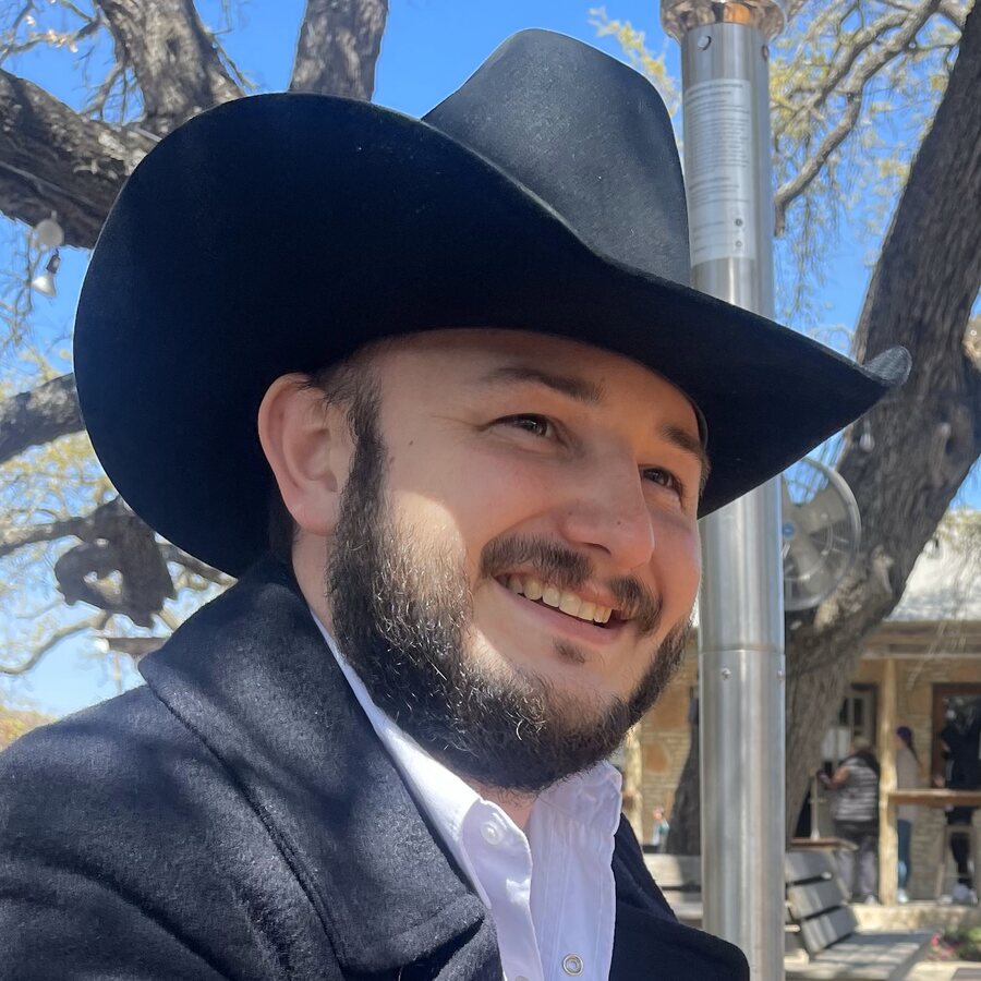 Coleton Pierson smiling in a black cowboy hat and dark coat, outdoors under live oak trees in Texas.